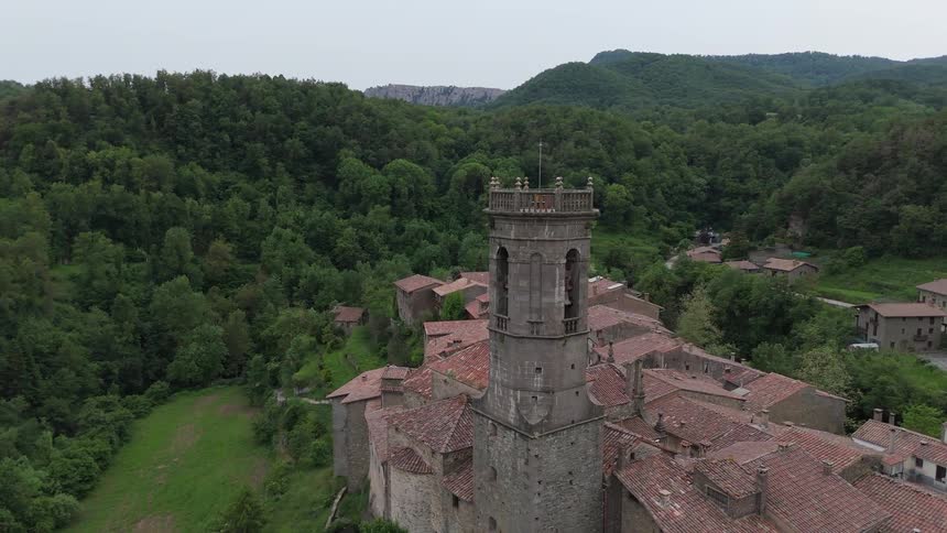 Historic village with a central tower surrounded by forested hills seen from above
