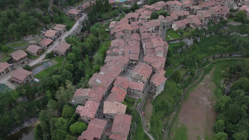 An aerial view pulls back to reveal a historic stone village with red tiled roofs perched on a lush forested ridge