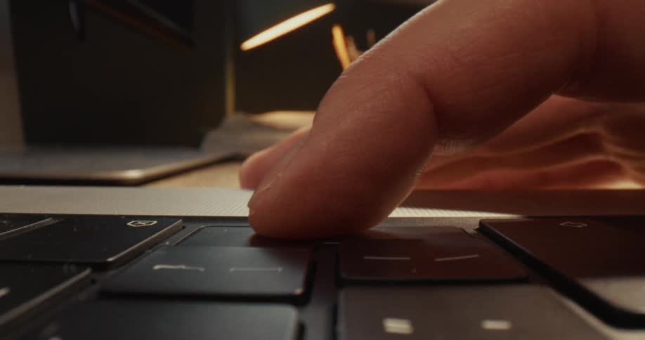 A finger presses the enter key on a laptop keyboard in a dimly lit workspace