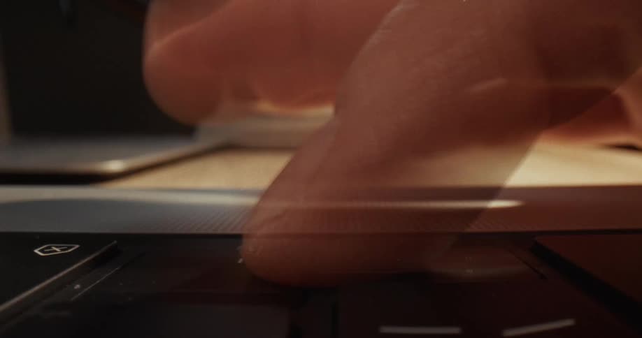 A person uses their index finger to press a key on a black laptop keyboard while sitting at a wooden desk under warm light