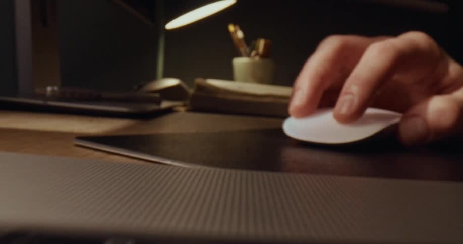 A person works at a wooden desk during the night using a white wireless mouse and a laptop