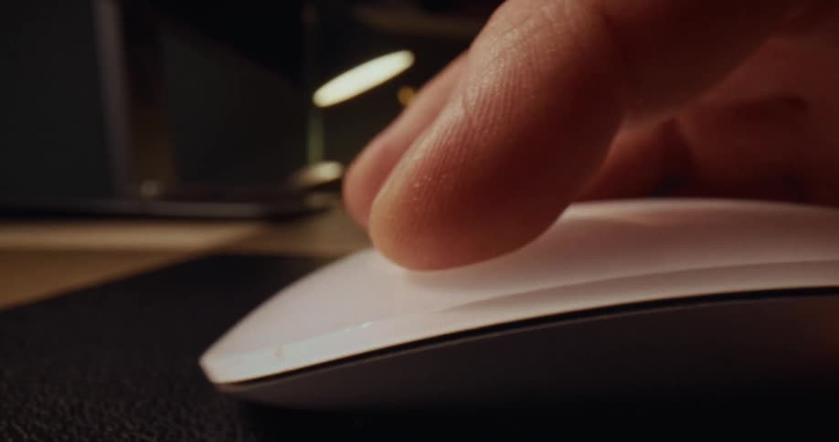 A person uses an index finger to click on a white computer mouse while sitting at a wooden desk in a dimly lit room with a lamp glowing in the background