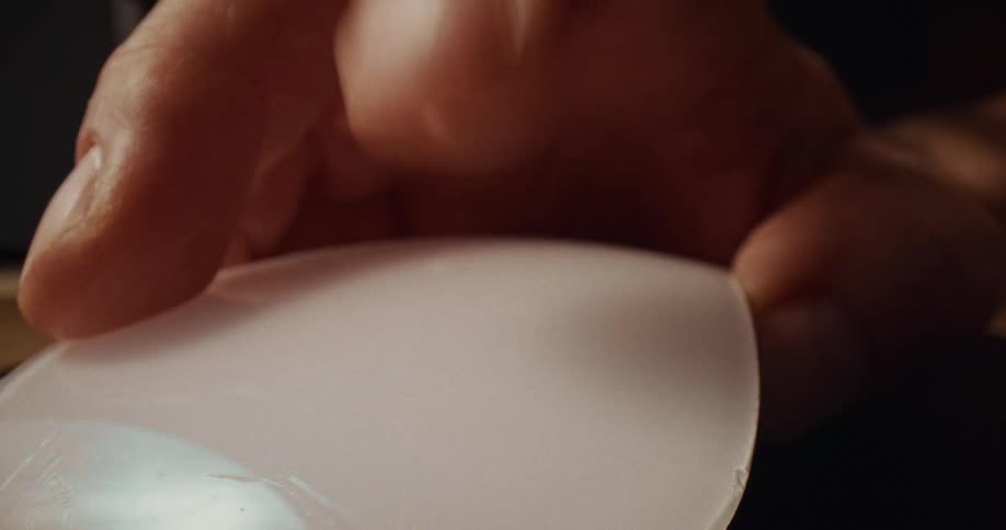 A closeup shot shows a persons fingers carefully holding and examining a translucent curved lens the individual appears to be inspecting the surface of the optical piece for clarity or imperfections under a soft light
