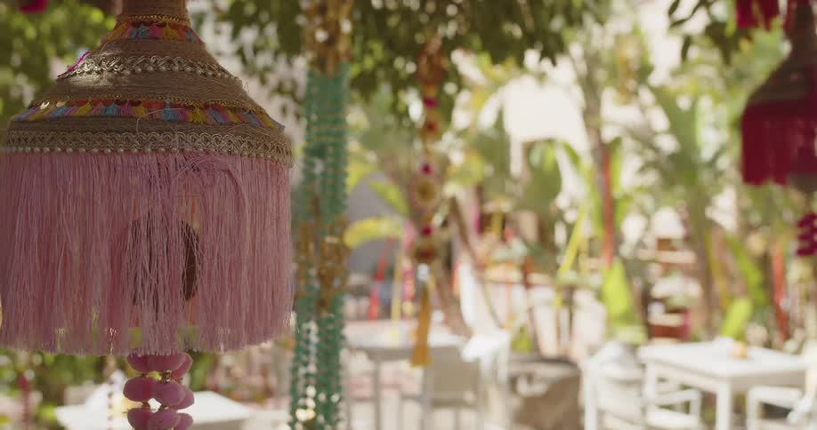 A close view of a pink decorative lantern with fringe hanging outdoors in front of a blurred patio area with white tables and green plants