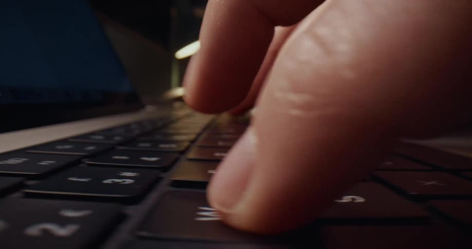 A man types quickly on a backlit laptop keyboard in a dimly lit room