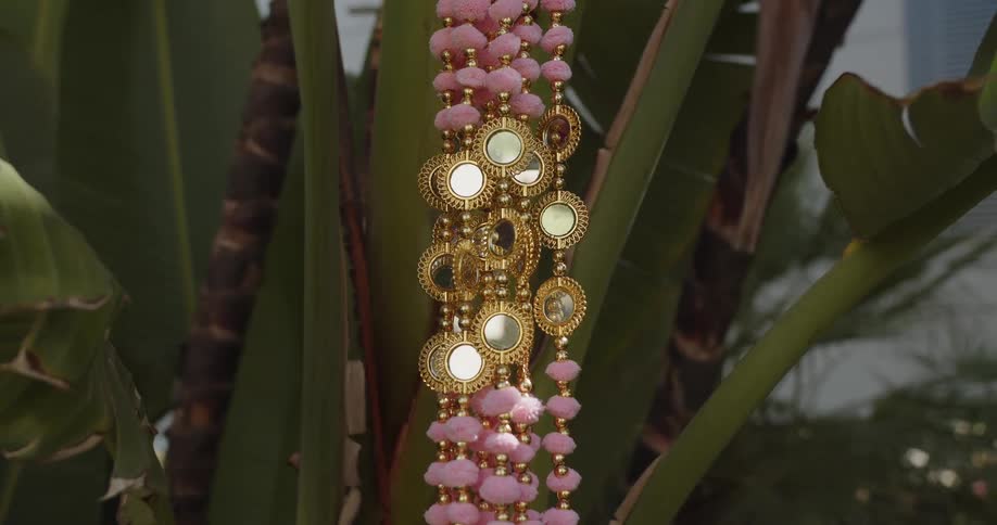 A close view of decorative pink bead strands with gold ornaments and small round mirrors hanging outdoors in front of large green tropical leaves on a bright day