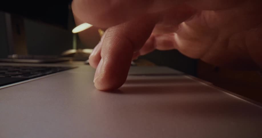A hand rests on a backlit keyboard and moves toward the trackpad of a silver laptopthe scene is shot from a low angle highlighting the texture of the fingertips and the warm ambient lighting in the background