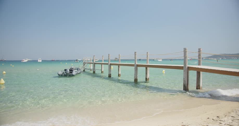 A grey boat is tied to a wooden pier that extends into clear turquoise water under a bright sky