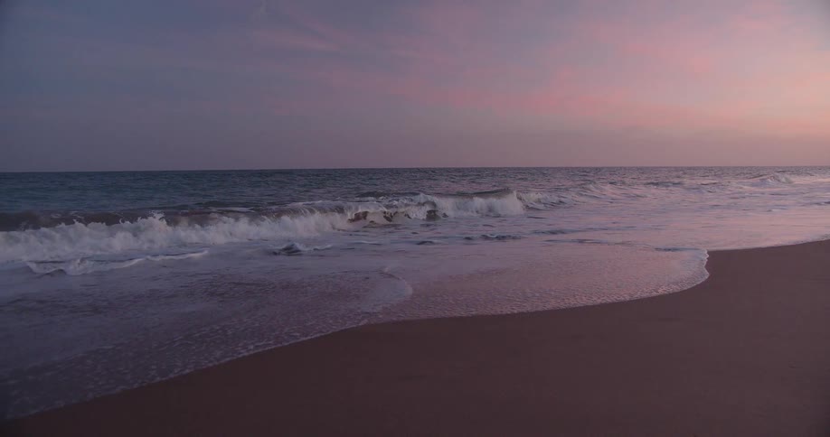 Gentle waves wash over a sandy beach beneath a pink and purple twilight sky