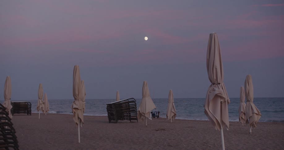 Wrapped beach umbrellas and chairs along the coast as waves roll in to shore and the moon glows in the sky