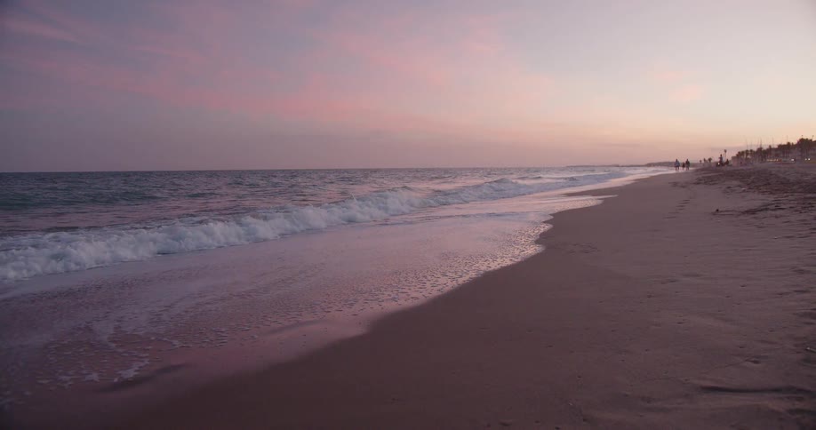 Gentle waves lap against a sandy shore under a soft pink and purple sunset sky in the distance several people walk along the waters edge as the tide slowly recedes