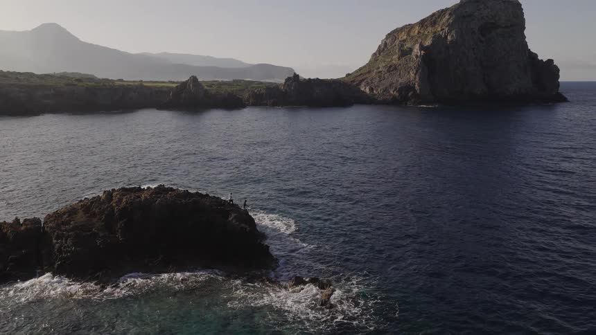 Waves are crashing against a small rocky island near a mountainous coast
