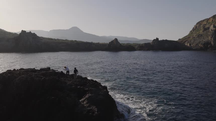 Two people stand on a dark rocky cliff overlooking a calm blue cove while the sun reflects off the water and mountains rise in the distance