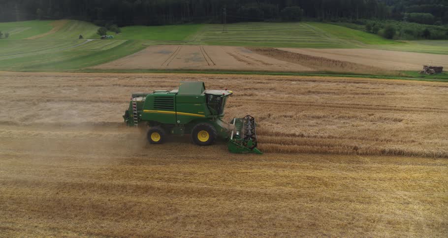 A green john deere combine harvester moves across a vast golden field cutting and threshing ripe wheat while leaving behind a trail of dust and straw against a backdrop of rolling green hills and a small wooden farm building