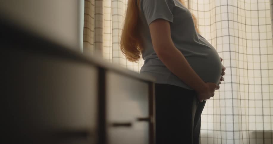 A pregnant woman stands in profile by a window gently cradling her baby bump with both hands she is wearing a lightcolored tshirt and dark pants framed against soft light filtering through a patterned curtain