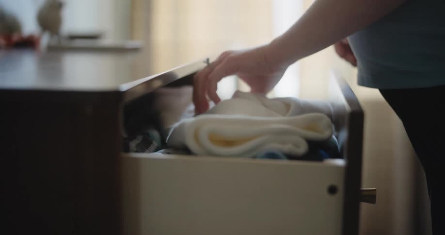A close up view shows a pregnant woman opening a wooden drawer and pulling out a small white garment