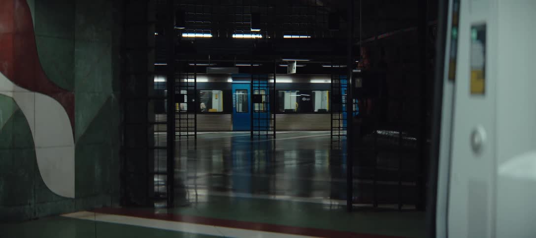 A blue and white subway train pulls into a dimly lit underground station platform viewed through a dark metal gate and concrete pillars
