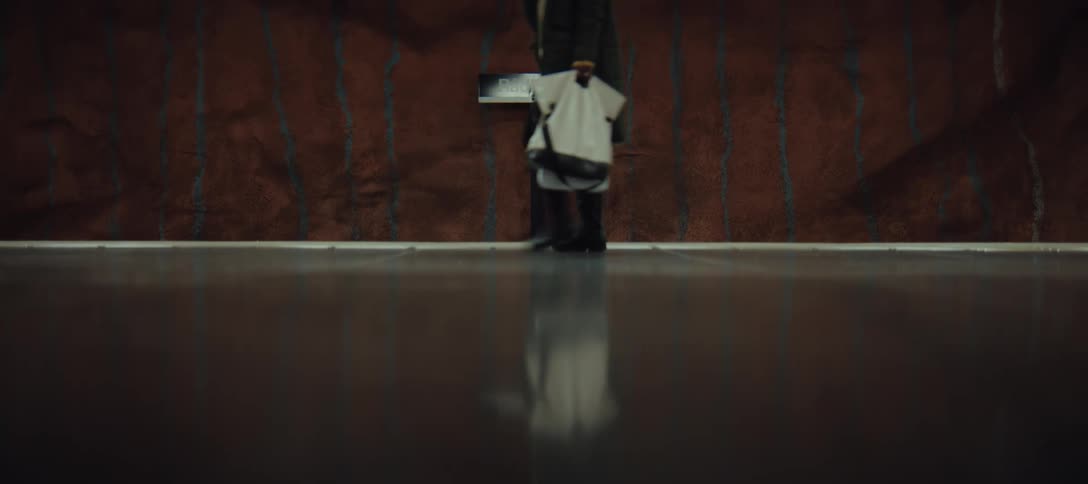 A person walking across a subway platform with their reflection visible on the polished floor the backdrop consists of a textured reddish rock wall and a station sign creating a moody cinematic atmosphere
