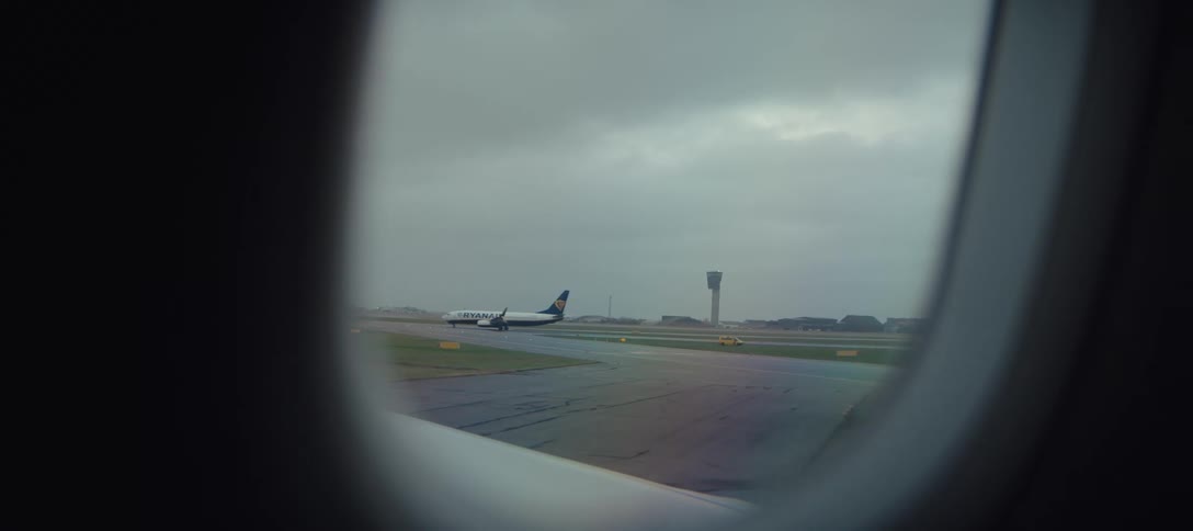 A view through an airplane window shows a jet taxiing on a wet runway under a cloudy sky