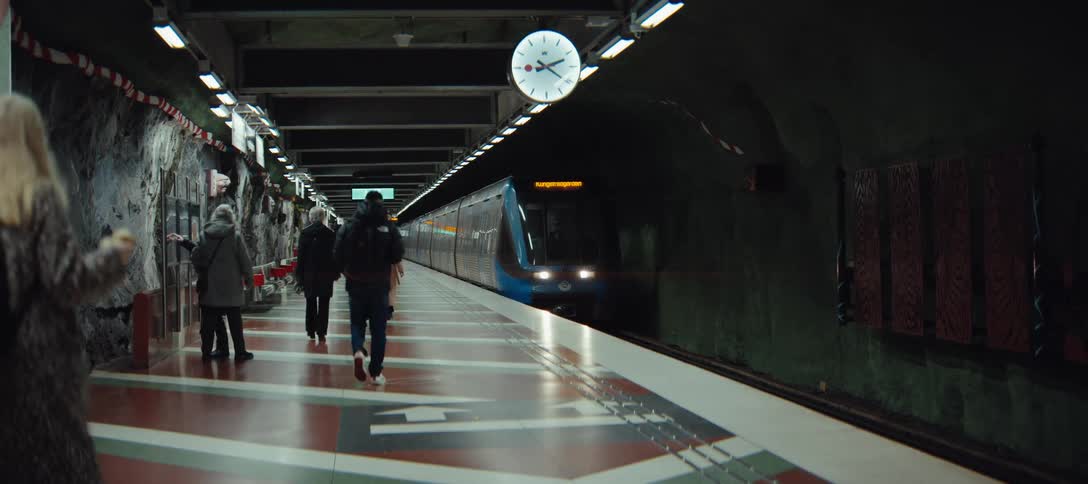People walk along a station platform as a blue train arrives from a dark tunnel under a large analog clock