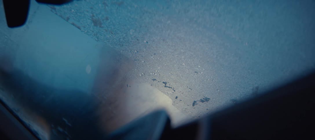 A close up view shows a person using a scraper to remove a layer of frost from the blue surface of a car windshield