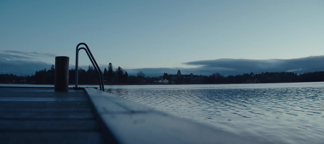 A low angle shot from a wooden pier looks across a calm lake toward a distant shoreline lined with trees and buildings under a dark blue morning sky with soft clouds resting on the horizon