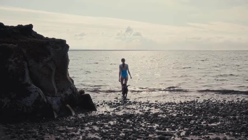 A woman in a blue swimsuit walks across a pebbly beach and enters the sea with her dog