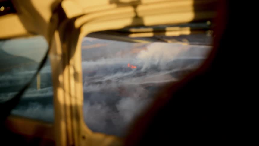 A view through an airplane window at sunset shows white smoke and glowing red lava from a volcanic eruption on the ground below
