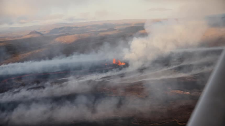 This aerial footage captures a volcanic eruption featuring a line of glowing lava fountains breaking through a desolate smoky landscape thick plumes of white and grey smoke drift across the dark terrain as the camera observes the activity from a moving