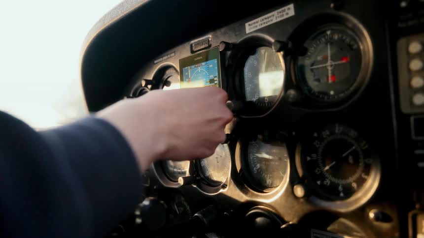 A pilots hand adjusts a digital flight instrument on a sunlit cockpit dashboard