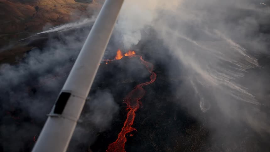 Glowing lava flow winding across the terrain with smoke seen from an aircraft