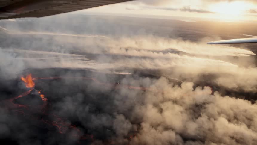 High above the earth a plane glides over a volcanic landscape where molten orange lava carves through the terrain beneath thick swirling plumes of white smoke and soft golden clouds at sunset
