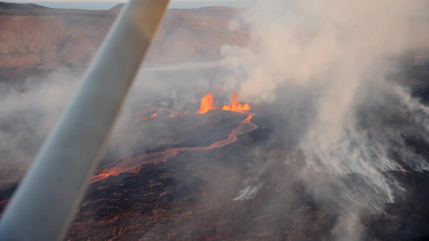 Lava flows over darkened terrain as thick smoke rises from active crater seen from aircraft
