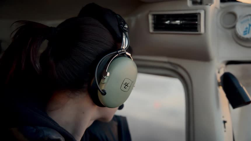 A woman wearing an aviation headset sits in a cockpit looking forward with a focused expression she then turns her head to look out the side window of the aircraft as it flies