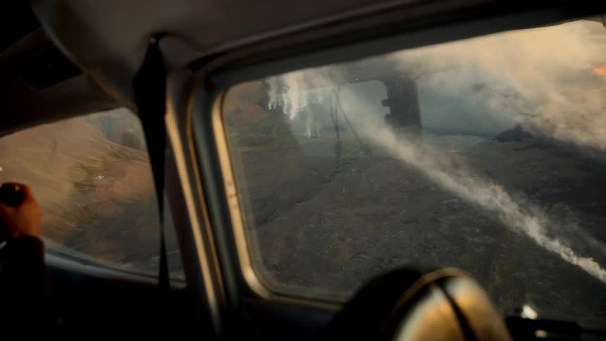 A passenger inside a small aircraft looks through a camera lens to photograph a rugged volcanic landscape from abovewisps of white smoke or steam drift across the dark rocky terrain as the plane glides over the earth during the golden hour