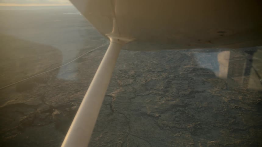 A person flies a light aircraft over a vast expanse of cracked desert earth during sunset as the golden light reflects off the wing strut and cockpit glass while the ground passes slowly below the plane
