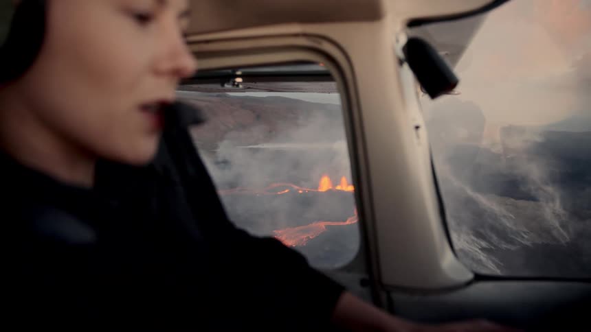 Woman wearing headphones inside a helicopter observing glowing lava through the side window
