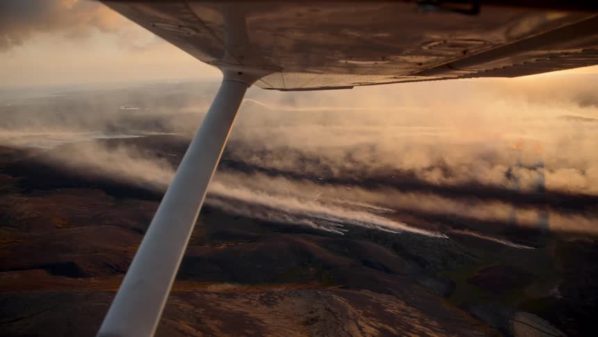 Smoke spreads over rugged terrain lit by the sun seen from aircraft