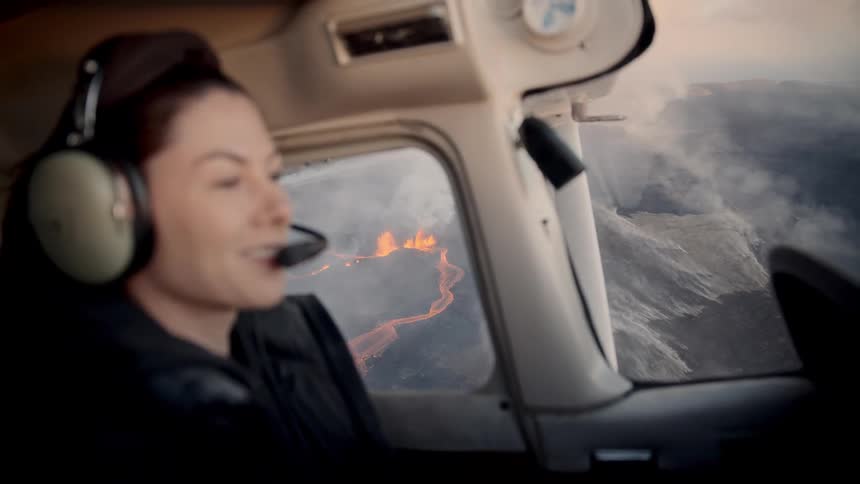 A woman wearing a headset watches a volcanic eruption through the window of a small airplane bright orange lava flows and smoke are visible across the dark landscape below the aircraft