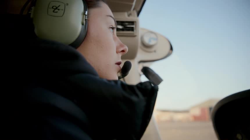 A female pilot wearing a professional headset sits in the cockpit of a small airplane and looks out the front window as the aircraft moves slowly across the runway toward the airport buildings under a clear blue sky during the day