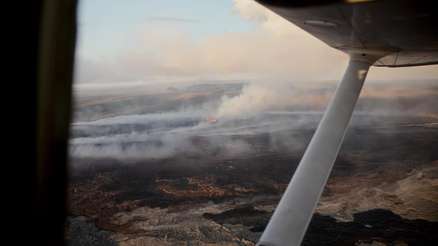 Incandescent lava emerges from fissures in volcanic terrain as seen from an aircraft in flight