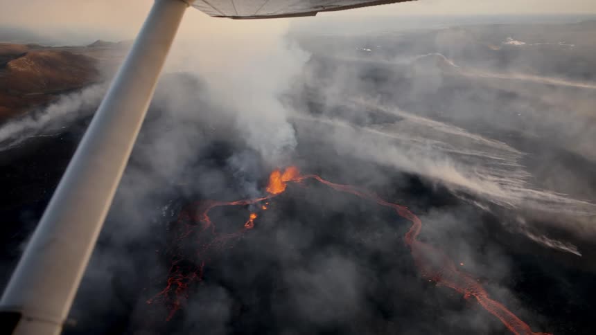 An aerial view from an airplane shows a volcanic eruption with glowing orange lava and thick white smoke rising from the dark earth below