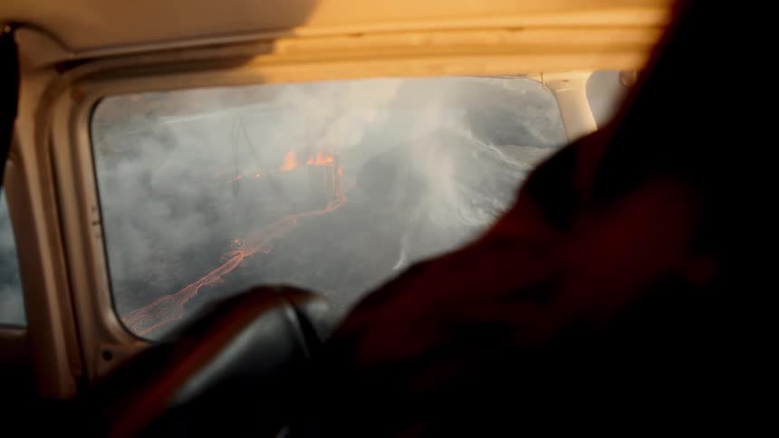A person sitting inside a small airplane looks through a side window at a volcanic eruption with orange lava and thick smoke on the ground below