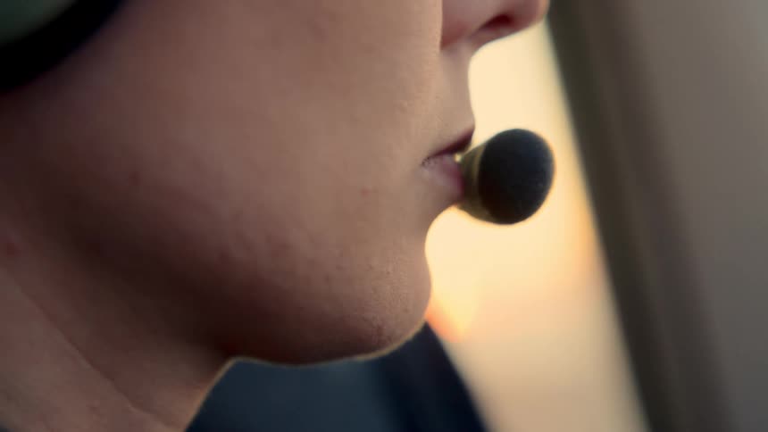 A closeup profile of a person wearing a professional aviation headset with a boom microphone positioned near their mouththe individual appears to be in a cockpit or aircraft cabin with warm golden light from a sunset or sunrise visible through the w