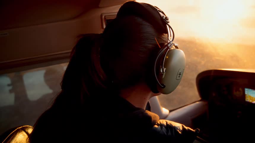 A woman wearing green headphones sits in the cockpit of a small airplane and looks out the side window during a flight at sunset