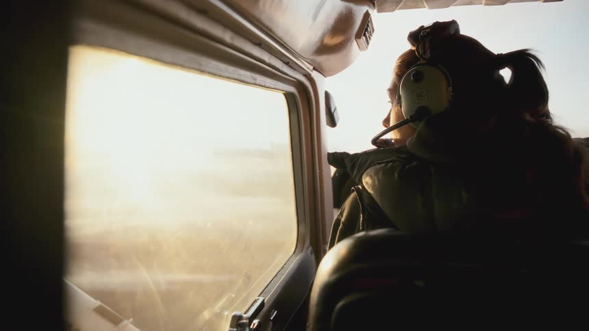 A woman wearing a headset gazes out the window of a small airplane at a sun drenched landscape