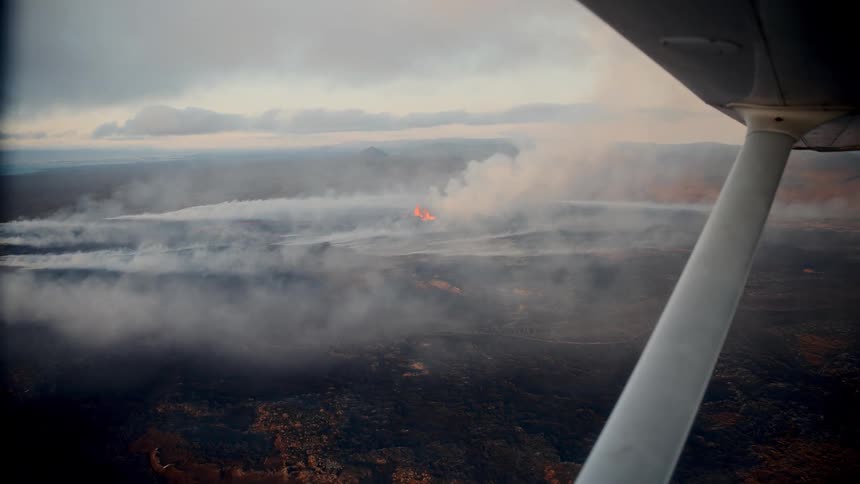 Aerial footage from a small plane shows a volcanic fissure erupting across a dark rocky landscape thick plumes of white smoke and orange lava fountains are visible beneath a cloudy overcast sky