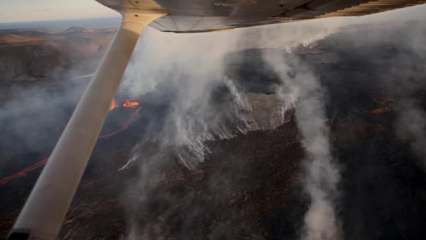 Aerial footage shows a plane flying over an active volcanic eruption with thick plumes of white smoke rising from the dark landscapeglowing orange lava flows are visible on the ground beneath the wing of the aircraft as it passes over the volcanic ter