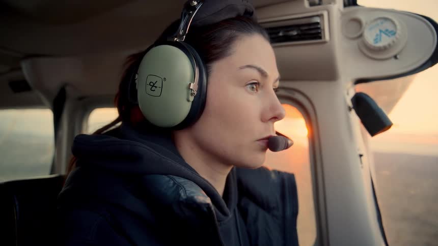 A woman wearing a flight headset gazes intently out of a cockpit window during a sunset flight the warm golden light of the horizon illuminates her face as she remains focused on the journey ahead