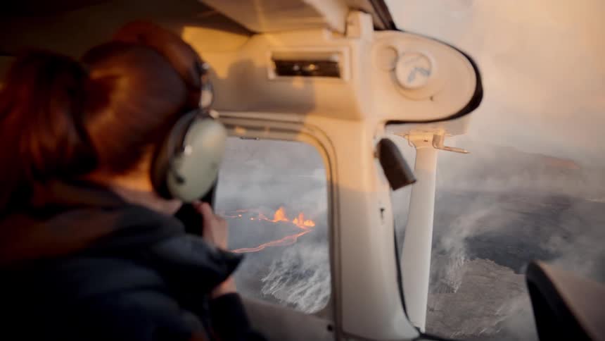 A pilot sitting inside a small airplane looks through a side window at a volcanic eruption with orange lava and thick smoke on the ground below taking a picture with her phone